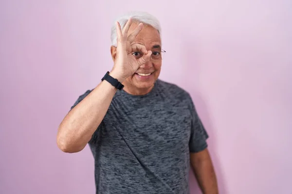 ピンクの背景の上に立っている灰色の髪を持つ中年男性は ハッピーフェイスで指を通して見て 手の笑顔でOkジェスチャーを行います — ストック写真