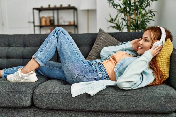 Genç beyaz kadın evdeki kanepede uzanıp müzik dinliyor.