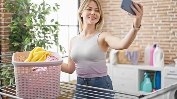 若い金髪の女性は 洗濯室で服のラインに掛かるスマートフォンでセルフィーを作ります — ストック写真