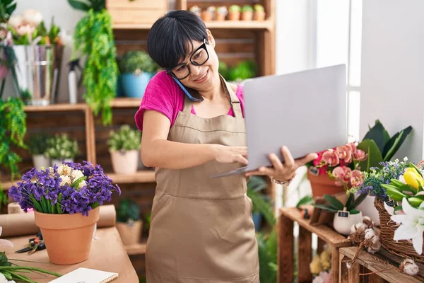 Middle age chinese woman florist talking on smartphone using laptop at flower shop