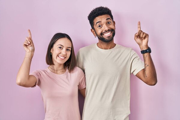 Young hispanic couple together over pink background smiling amazed and surprised and pointing up with fingers and raised arms. 