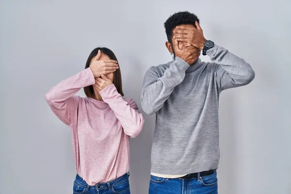 Young hispanic couple standing together covering eyes and mouth with hands, surprised and shocked. hiding emotion