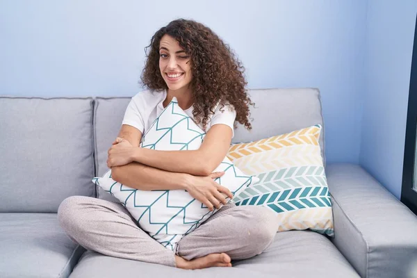 Hispanic woman with curly hair sitting on the sofa at home winking looking at the camera with sexy expression, cheerful and happy face. 