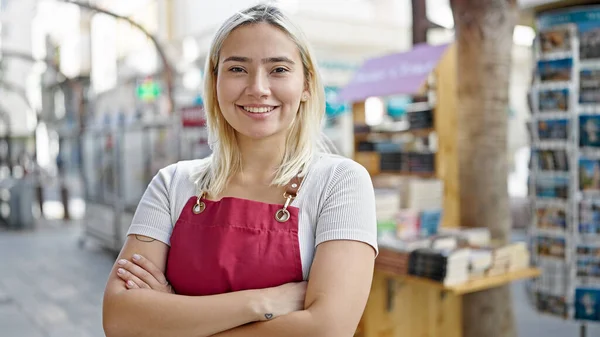 Önlük giymiş genç, İspanyol bir kadın gazete bayiinde kollarını kavuşturup el kol hareketi yapıyor.