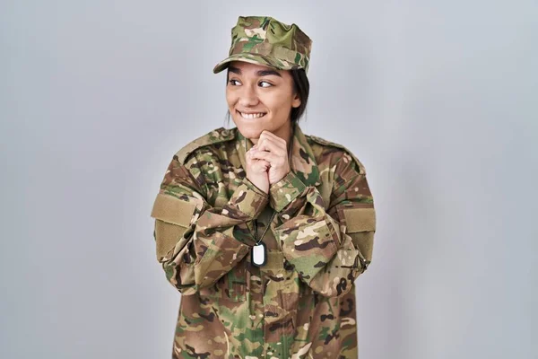 Young south asian woman wearing camouflage army uniform laughing nervous and excited with hands on chin looking to the side