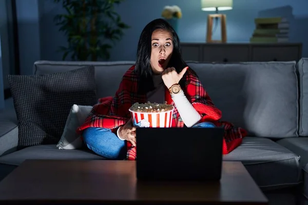 Hispanic Woman Eating Popcorn Watching Movie Sofa Surprised Pointing Hand — стоковое фото
