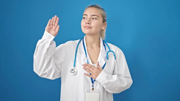 年轻美丽的惊慌失措的女医生 手托着胸膛在孤零零的蓝色背景下宣誓 — 图库照片