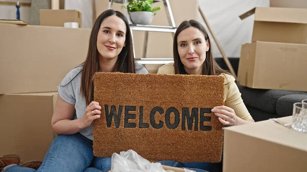 Two women holding welcome doormat sitting on floor at new home