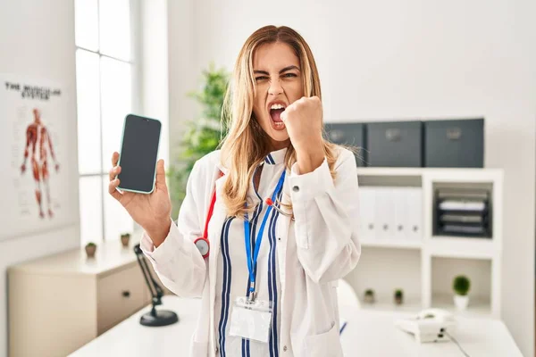Young Blonde Doctor Woman Working Clinic Showing Smartphone Screen Annoyed — Stockfoto