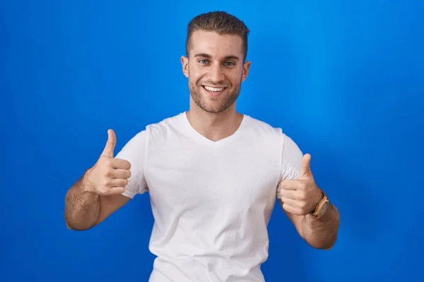 年轻的高加索男子站在蓝色背景的成功的标志上 用手做着积极的手势 竖起大拇指 微笑着 快乐地 快乐的表达和胜利的姿态 — 图库照片