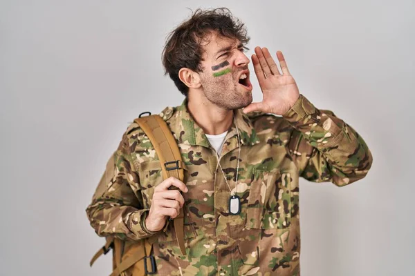 Hispanic young man wearing camouflage army uniform shouting and screaming loud to side with hand on mouth. communication concept.