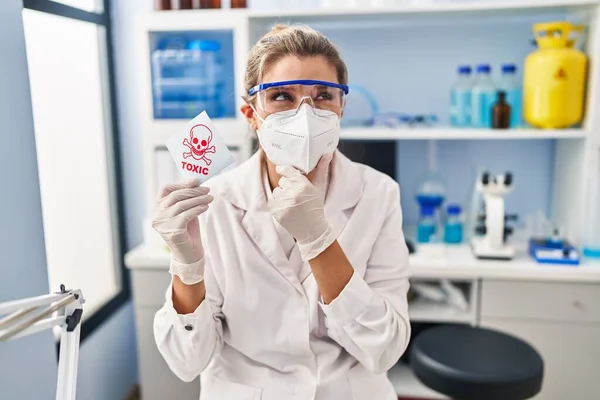 在科学家实验室工作的年轻女子拿着有毒横幅严肃的脸 手托着下巴思考着问题 思考着困惑的想法 — 图库照片