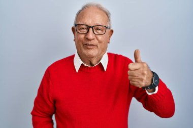 Senior man with grey hair standing over isolated background doing happy thumbs up gesture with hand. approving expression looking at the camera showing success. 