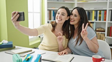 Masada oturup ders çalışan iki kadın kütüphane üniversitesinde akıllı telefondan selfie çekiyor.