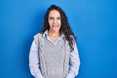 Young brunette woman standing over blue background sticking tongue out happy with funny expression. emotion concept. 