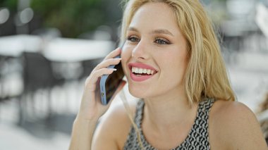 Genç sarışın kadın kahve dükkanının terasında oturmuş akıllı telefondan konuşuyor.