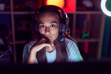 Young beautiful hispanic woman streamer sitting on table with serious expression at gaming room