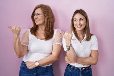 İspanyol anne ve kızı pembe arka planda günlük beyaz tişört giyip gülümsüyor ve başparmağıyla yan tarafa bakıyor.. 