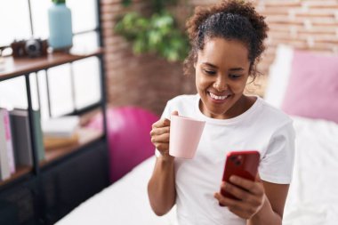 African american woman using smartphone drinking coffee at bedroom