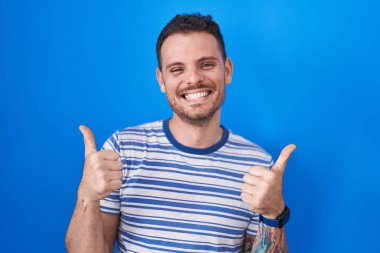 Young hispanic man standing over blue background success sign doing positive gesture with hand, thumbs up smiling and happy. cheerful expression and winner gesture. 