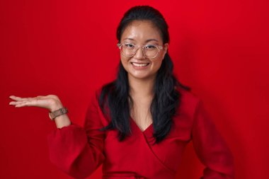 Asyalı genç bir kadın kırmızı arka planda durup gülümsüyor. Neşeli bir şekilde sunuyor ve avuç içiyle kameraya bakıyor.. 