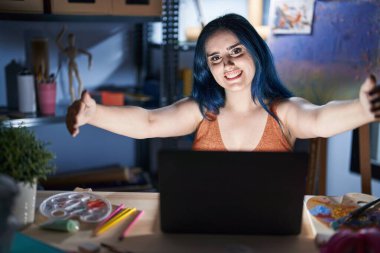 Mavi saçlı genç modern bir kız geceleri dizüstü bilgisayarla resim stüdyosunda oturuyor ve kameraya sarılıp gülümsüyor. Mutluluğu kucaklayan neşeli bir ifade.. 