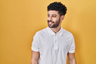 Arap adam sarı arka planda duruyor. Yüzünde gülümseme ve doğal ifadeyle yan yana bakıyor. Kendinden emin gülen. 