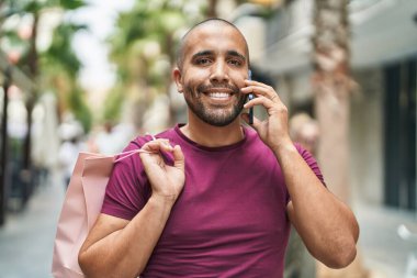 Young latin man talking on smartphone holding shopping bag at street