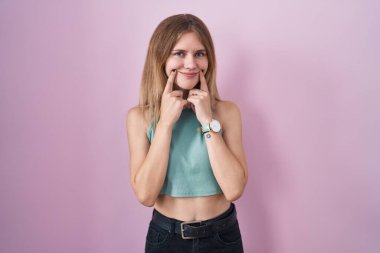 Pembe arka planda duran sarışın beyaz kadın ağzı açık gülümsüyor, parmakları işaret ediyor ve neşeli bir gülümseme sergiliyor. 