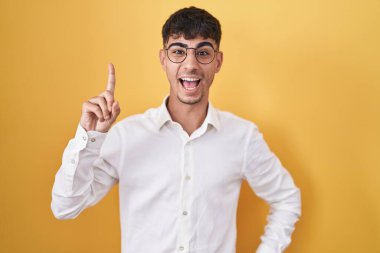 Young hispanic man standing over yellow background pointing finger up with successful idea. exited and happy. number one. 