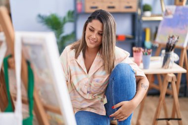 Genç İspanyol kadın sanatçı, resim stüdyosunda yerde otururken kendine güvenerek gülümsüyor.