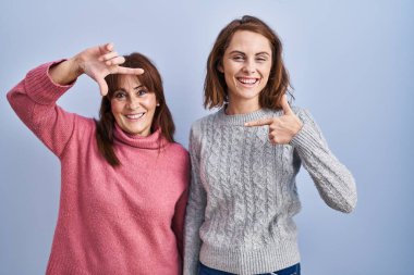 Anne ve kızı mavi arka planda gülümsüyor ve mutlu yüzlü eller ve parmaklarla çerçeve yapıyorlar. yaratıcılık ve fotoğrafçılık kavramı. 