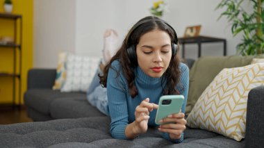 Akıllı telefon kullanan, kulaklık takmış, evdeki kanepede uzanmış genç, güzel İspanyol bir kadın.