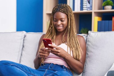 Evdeki koltukta oturan Afro-Amerikan kadın akıllı telefon kullanıyor