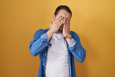 İspanyol adam sarı arka planda duruyor. Yorgunluk ve baş ağrısı için gözlerini ovuşturuyor. Uykulu ve yorgun bir ifade. görme sorunu 