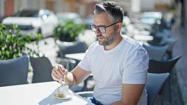 Masada oturan gri saçlı adam kahve dükkanının terasında kahveyi karıştırıyor.