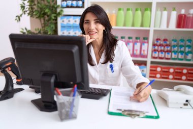 Middle age hispanic woman pharmacist writing on document using computer at pharmacy