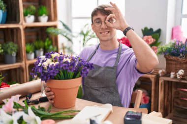 Beyaz, sarışın bir adam çiçekçide çalışıyor. El hareketleriyle gülümsüyor ve mutlu yüzüyle parmaklarının arasından bakıyor.. 