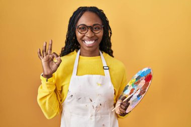Afrikalı bir kadın ressam paleti tutuyor. Gülümsüyor. El ve parmaklarıyla tamam işareti yapıyor. başarılı ifade. 
