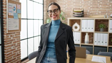 Genç, güzel, İspanyol bir iş kadını ofiste kendinden emin gülümsüyor.