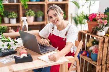 Young blonde woman florist using laptop writing on notebook at flower shop