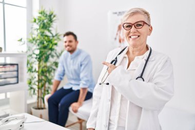 Tıbbi klinikte üniforma ve steteskop giyen kadın doktor mutlu bir şekilde elini ve parmağını göstererek gülümsüyor. 