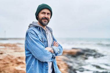 Kel genç adam sahilde kollarını kavuşturmuş el kol hareketi yapıyor.