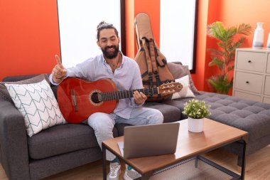 Sakallı İspanyol bir adam evde klasik gitar çalıyor. Gülümsüyor, eli ve parmağı kenarda mutlu bir şekilde işaret ediyor. 