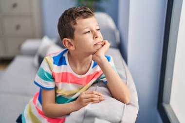 Evde ciddi bir ifadeyle koltukta oturan sarışın çocuk.