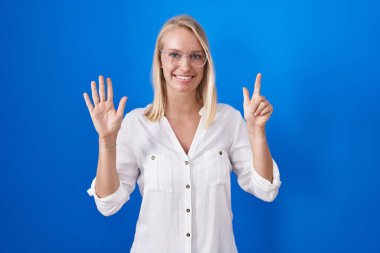 Young caucasian woman standing over blue background showing and pointing up with fingers number seven while smiling confident and happy. 