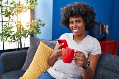 Afro-Amerikalı kadın akıllı telefon kullanıyor evde kahve içiyor.