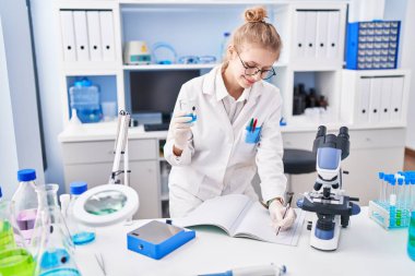 Young blonde woman scientist writing on notebook holding test tube at laboratory