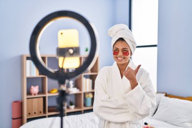 Koyu renk saçlı İspanyol bir kadın akıllı telefonuyla güzellik dersi kaydediyor mutlu ve olumlu gülümsüyor, başparmağıyla mükemmel ve onay işareti yapıyor. 