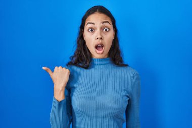 Mavi izole edilmiş arka planda dikilen Brezilyalı genç bir kadın elleri yana dönük, ağzı açık şaşkın bir ifadeyle şaşırdı.. 